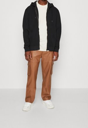 Mężczyzna ubrany w czarną bluzę z kapturem zapinaną na zamek, białą koszulkę, brązowe spodnie cargo i białe sneakersy, stojący na jednolitym tle.