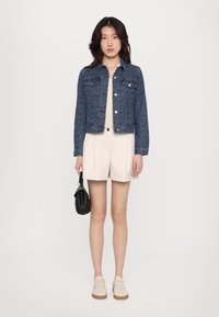 Dunkelblaue Jeansjacke mit strukturiertem Design, hellbeiges Oberteil, cremfarbene Shorts und beigen Sneakern. Schwarze Handtasche in der rechten Hand gehalten.