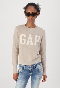 Light beige, knitted sweater with white "GAP" logo on the front. Paired with distressed blue jeans. Model wears black sunglasses.