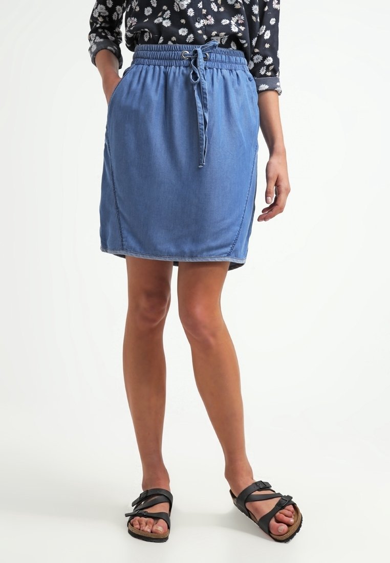 Femme portant une jupe en denim bleue avec une taille à cordon, associée à des sandales noires et une chemise noire à manches longues avec motif floral, debout les mains dans les poches.