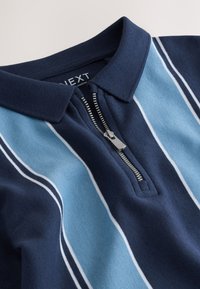 Polo bleu marine avec des rayures verticales bleu clair et blanches, doté d'une courte fermeture éclair argentée et d'un col plié.