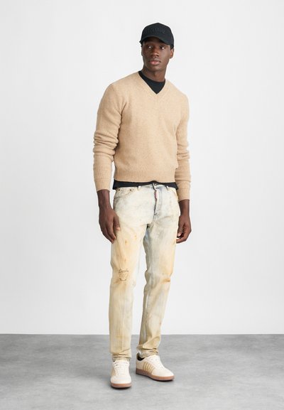 Beige gestrickter Pullover, schwarzes Hemd darunter, hell ausgebleichte Jeans mit distressed Details und weiße Sneaker mit beigen Akzenten. Schwarze Mütze.
