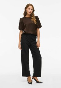 Een vrouw staat gekleed in een donkergroene blouse met pofmouwen, zwarte hoge taille wijd uitlopende broek en zwarte puntige hakken met gouden schakels.