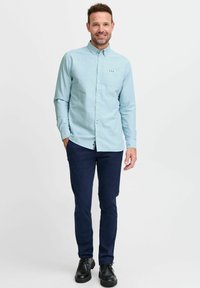Camicetta blu chiaro con taschino sul petto, abbinata a pantaloni blu navy e scarpe nere. La camicia presenta un piccolo logo.