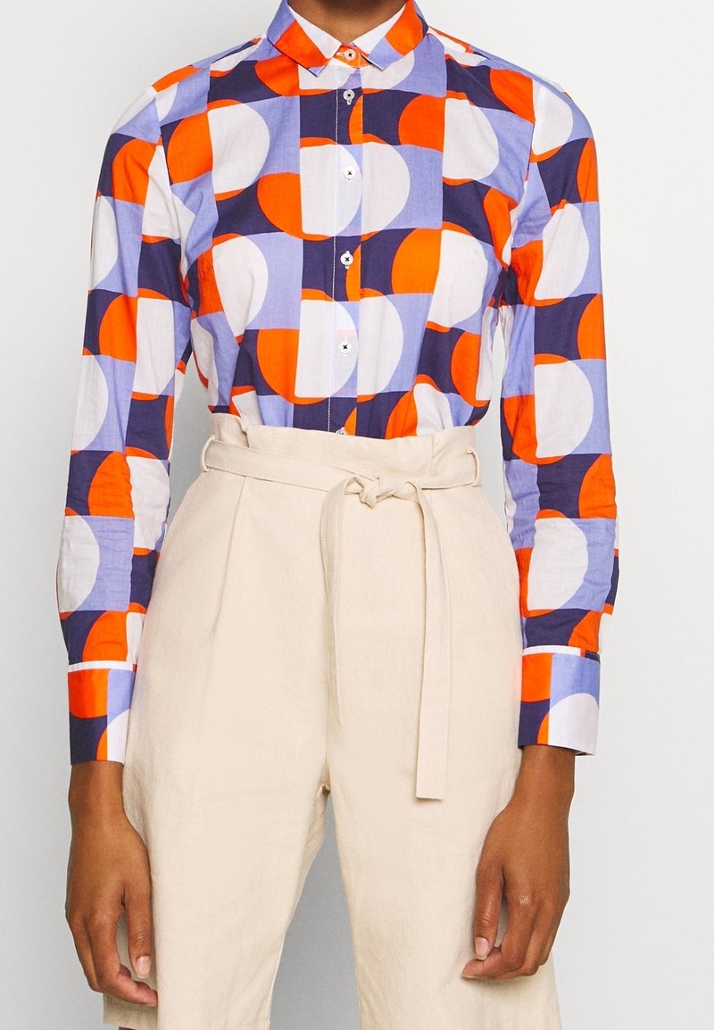 Chemise à col avec motif géométrique orange, violet et blanc, associée à un pantalon beige taille haute noué avec une ceinture en tissu.