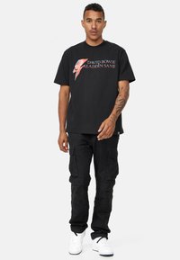 Svart bomull t-shirt med ett grafiskt tryck av en blixt och texten "David Bowie Aladdin Sane" i blått och rött. Bärs tillsammans med svarta cargobyxor.
