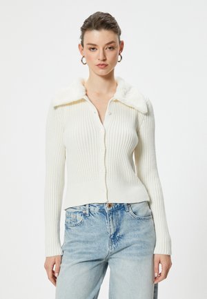 Cardigan bianco a coste con colletto in morbido pelliccia sintetica, dotato di chiusura con bottoni e maniche aderenti, abbinato a jeans in denim azzurro chiaro.