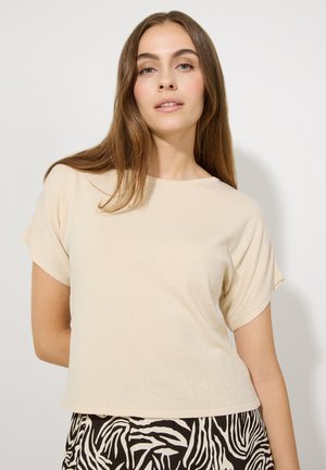 Beige Kurzarmoberteil aus einem weichen, strukturierten Stoff. Mit Rundhalsausschnitt und lockerer Passform, kombiniert mit einem gemusterten schwarz-weißen Rock.