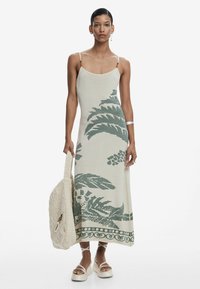 Megztas maxi suknelė off-white spalvos su žaliais botaniniais raštais, turinti plonas petnešas ir tiesią siluetą. Derinama su nertine rankine.