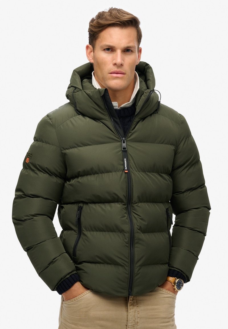 Uomo che indossa un giubbotto imbottito verde oliva con tasche zip, pantaloni beige, maglione scuro a maglia, colletto della camicia bianco e un orologio da polso.