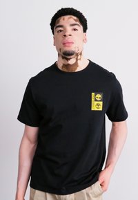 Zwarte katoenen t-shirt met een rechthoekige gele logopatch op de linkerborst, ronde halslijn en korte mouwen. Glad oppervlak, ontspannen pasvorm.