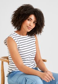 Woman with curly hair wearing a white sleeveless top with black stripes, sitting on a wooden chair with hands resting on her lap.