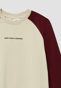 Crèmekleurige sweatshirt met bordeauxrode raglanmouwen. Voorzien van eenvoudige geborduurde tekst "SLVR STUDIOS | DEPARTEMENT" in bordeaux. Zachte textuur.
