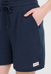 Close-up of navy textured shorts with drawstring waist, hand in right pocket, and small "oxmo" label near hem.