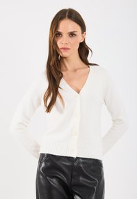 Krémové, priliehavé kardigán s výstrihom do V, dlhými rukávmi a piatimi gombíkmi, vyrobené z mäkkého, textúrovaného materiálu. Rímský lem.