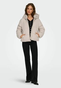 Giacca a vento beige chiaro con cappuccio, dotata di chiusura con zip e due tasche frontali, indossata sopra una maglietta nera e abbinata a pantaloni neri a zampa.