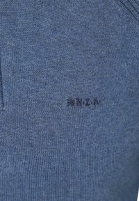 Blauwe gebreide stof met kleine, geborduurde donkere tekst "NZA" nabij het midden.