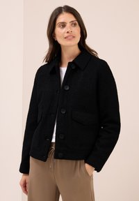 Donna che indossa una giacca in pile nera con bottoni su una maglietta bianca e pantaloni beige, in piedi con una mano in tasca e guardando di lato.