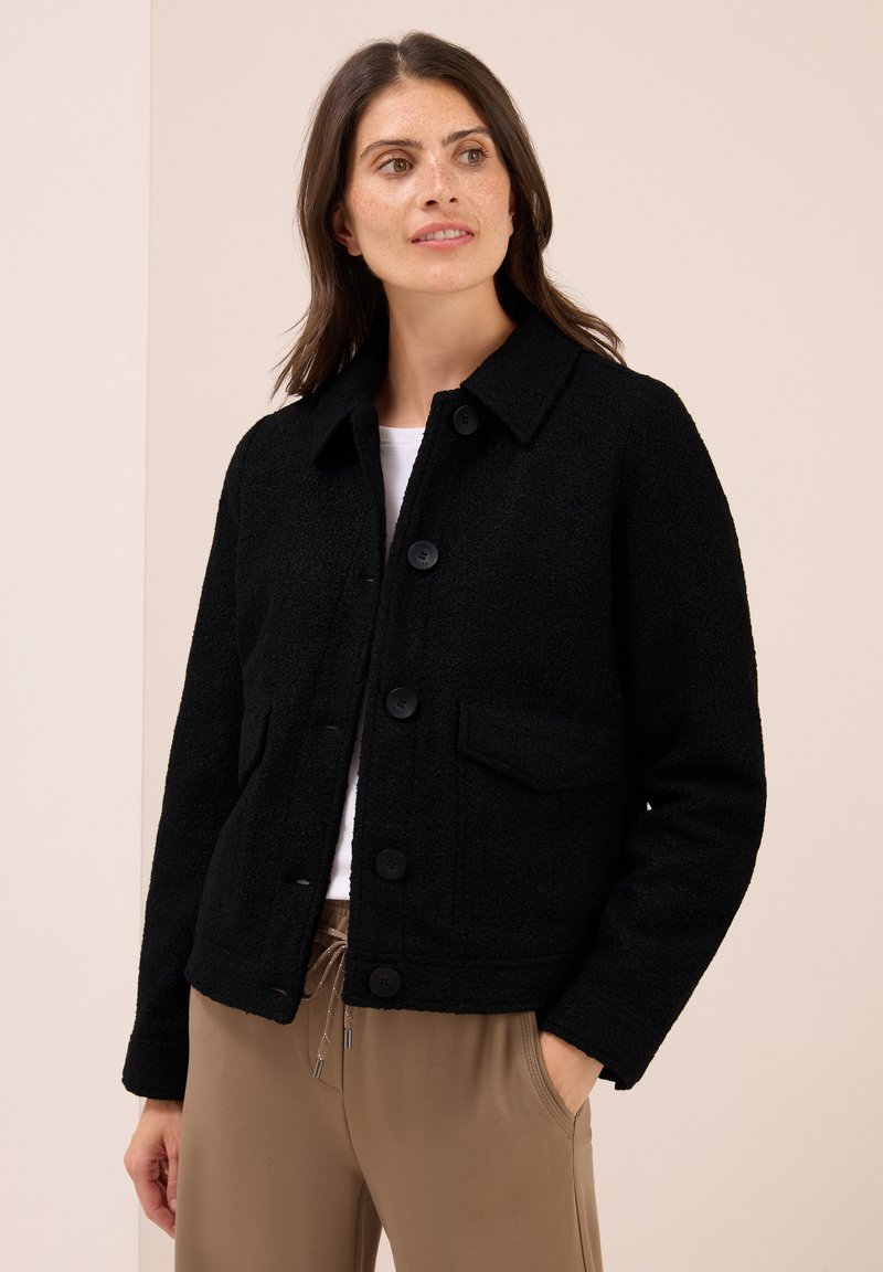 Kvinde iført sort fleecejakke med knapper over hvid top og beige bukser, stående med den ene hånd i lommen og kiggende til siden.