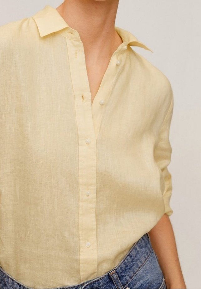 Chemise en lin jaune clair boutonnée, portée rentrée dans un jean en denim bleu, avec les manches retroussées de manière décontractée.