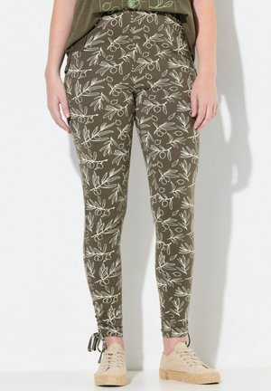 Olivgrüne Leggings mit einem weißen Blattmuster, die ein figurnahes Design und verstellbare Bindebänder an den Knöcheln aufweisen, kombiniert mit hellen Schuhen.
