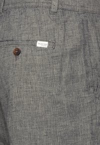Grå teksturerte bukser med herringbone-mønster, med en brun knapp og et lite merke som sier "SELECTED HOMME."