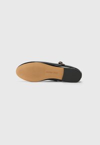 Melnā lakota ādas loafer kurpe ar brūnu zoli un melnu papēdi, kurai ir zelta sprādze un "Steve Madden" zīmols uz zoles.