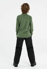 Maglione in pile verde con colletto alto, abbinato a pantaloni da neve neri isolati, visti da dietro. La morbidezza è evidente nel tessuto.