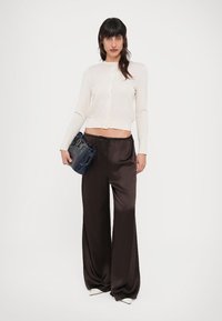 Ανοιχτό μπεζ κοντό ζακέτα, καφέ σατέν παντελόνι με φαρδύ παντελόνι και μια σκούρα μπλε τσάντα με δομημένο σχέδιο. Το μοντέλο φοράει λευκά παπούτσια με τακούνι.