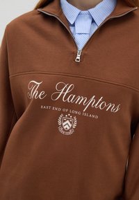 Felpa marrone a collo mezzo zip con la scritta bianca "The Hamptons East End of Long Island" e un blasone sopra una camicia collare a righe blu.