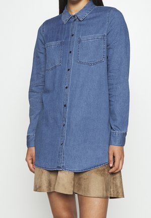 Denim overhemd in medium blauw, lange mouwen, knoopsluiting aan de voorkant, twee borstzakken. De stof is glad met een lichte glans, gecombineerd met een beige rok.
