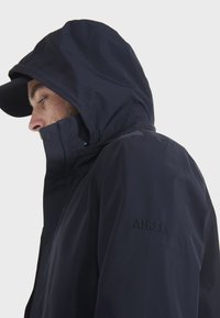 Navy wasserfeste Jacke mit Kapuze. Verfügt über einen hohen Kragen, einen Reißverschluss vorne und ein gesticktes Logo am Arm. Glatte Textur, keine sichtbaren Muster.