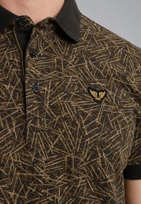 Kortærmet polo shirt i mørkebrun med et geometrisk mønster af beige og lysebrune linjer. Har en sort krave og logo-patch.