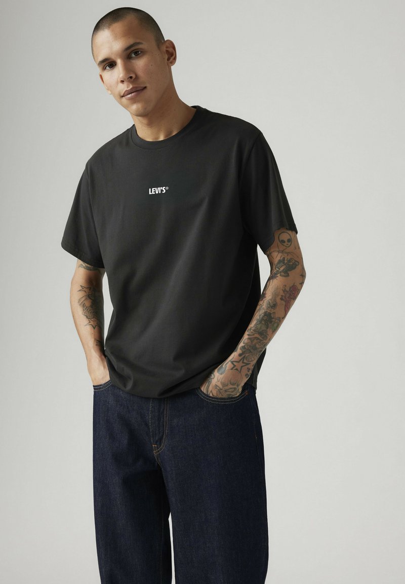 Joven con la cabeza rapada, viste una camiseta negra de Levi's y vaqueros azul oscuro, manos en los bolsillos, tatuajes visibles en los brazos, fondo gris neutro.
