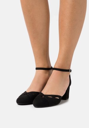 Black suede heels with a block heel, open toe, and ankle strap featuring cut-out accents across the upper. Smooth texture overall.