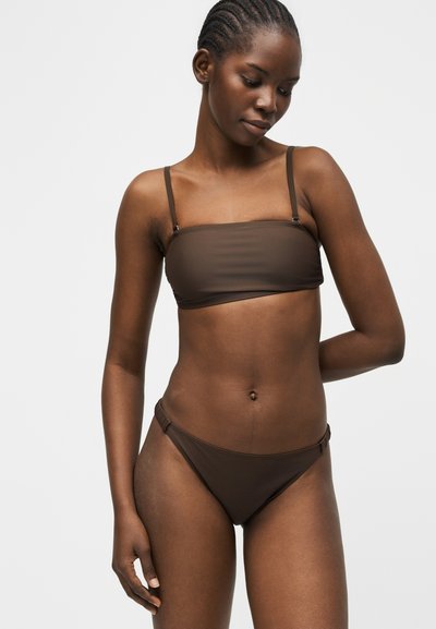 Γυναίκα με πλεγμένα μαλλιά που φοράει μπαντό μπικίνι σε σκούρο καφέ χρώμα και αντιστοιχά κιλοτάκια μπικίνι, με φόντο απλό και ανοιχτόχρωμο.