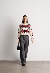 Krém színű kötött pulóver piros és fekete geometriai mintákkal, szürke csíkos nadrággal és egy kis fekete táskával párosítva.