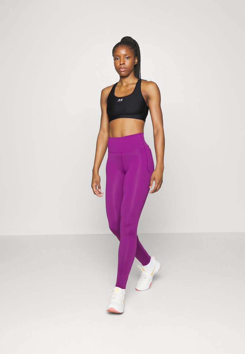 Sostén deportivo negro con un logo, combinado con leggings morados de tiro alto y lisos. El modelo lleva zapatillas deportivas blancas con acentos en tonos pastel.