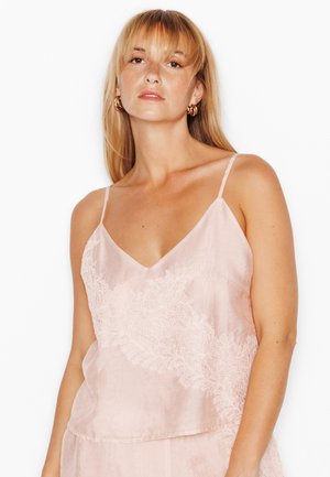Femme aux longs cheveux blonds portant des boucles d'oreilles créoles dorées et un haut sans manches rose pâle avec des détails en dentelle sur fond blanc.