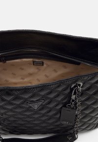 Czarna pikowana torba na ramię o teksturowanym wykończeniu, zamykana na zamek, beżowe wnętrze z wzorem oraz pasek zdobiony łańcuszkiem. Widoczne logo marki.