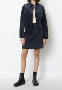 Tmavě námořnická manšestrová bunda s dvěma náprsními kapsami v kombinaci se stejnou sukní; modelka má na sobě černé kotníkové boty; minimalistický design a textura.
