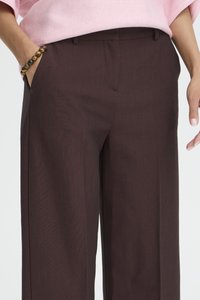 Frau trägt dunkelbraune, weit geschnittene Hosen und ein rosa Oberteil, mit einer Hand in der Tasche und einem Perlenarmband am Handgelenk.