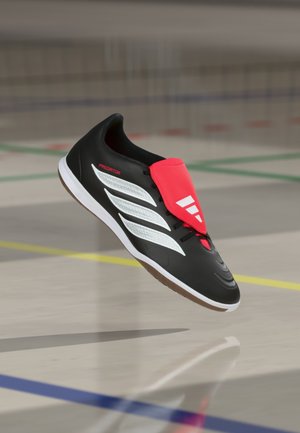 Chaussure de football indoor noire avec des bandes blanches, protection de lacet rouge affichant un logo, flottant au-dessus d’un sol de gymnase avec des lignes de terrain colorées.