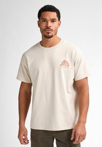 Beige katoenen t-shirt met een ronde hals, korte mouwen en een palmprint in lichtbruin op de linkerkant van de borst.