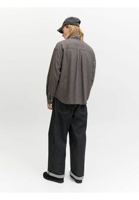 Person iført en ternet langærmet skjorte, mørke vidde jeans med opretstående ben, sorte sko og en mørk baseballkasket, vendende væk mod en hvid baggrund.