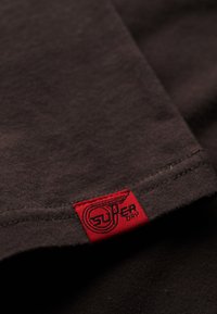 Camiseta de algodón marrón oscuro con una etiqueta roja que presenta el logo de "Superdry" y un gráfico de alas. Textura suave, cuello redondeado y dobladillo cosido.