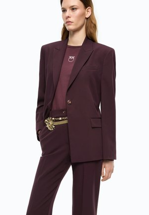 Donna che indossa un completo sartoriale viola scuro con camicia abbinata e cintura a catena d’oro con un grande ornamento a forma di fiore.