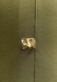 Tkanina w oliwkowym kolorze o gładkiej strukturze posiada metaliczny złoty akcent w postaci kokardy przymocowanej w szwie. Widoczne są detale przeszycia.