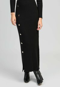 Jupe midi noire en tricot côtelé avec une fente sur le côté et six boutons métalliques sur le devant. Portée avec des bottines noires.