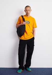 Borsa nera trapuntata portata a spalla, t-shirt arancione con logo verde, pantaloni neri e scarpe turchesi con strisce rosse.